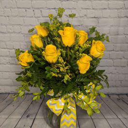 Arrangement of yellow roses with lush foliage in a clear vase with a yellow chevron ribbon