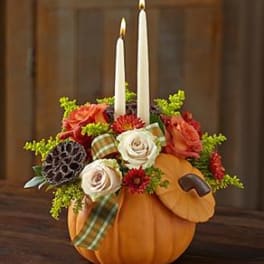 Autumn centerpiece of orange and cream flowers with two taper candles in a pumpkin-shaped container