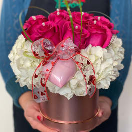 Pink roses and white flowers arranged in a round gift box with a ribbon and heart ornament.
