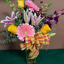 Mixed bouquet of pink gerbera daisies, yellow roses, lilies, and orchids in a glass vase