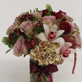 Bouquet of dusty pink roses, red roses, orchids, and hydrangea in a glass vase with a burgundy ribbon.