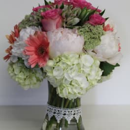 Round bouquet of pink roses, white hydrangeas, and coral gerberas in a glass vase with lace trim