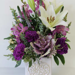 Tall arrangement of white lilies and purple flowers in a white ceramic cube vase with embossed heart.