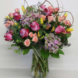 Tall mixed arrangement of pink roses, peach spray roses, yellow tulips, and purple hydrangeas in a glass vase