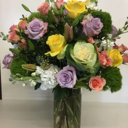 Tall mixed arrangement of yellow, lavender, and peach roses with lilies in a clear glass vase