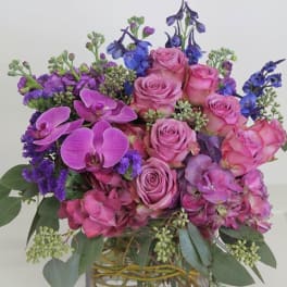 Compact arrangement of pink roses, orchids, and hydrangeas with purple and blue accents in a clear glass cube vase