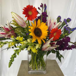 Mixed bouquet with sunflowers, gerbera daisies, lilies, and purple blooms in a glass vase