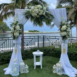 White floral wedding arch with draped fabric and lanterns by the water