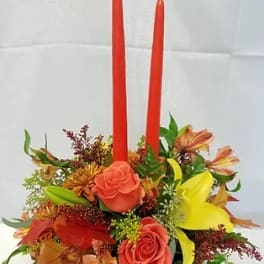 Floral centerpiece with two red taper candles and mixed orange, yellow, and pink blooms