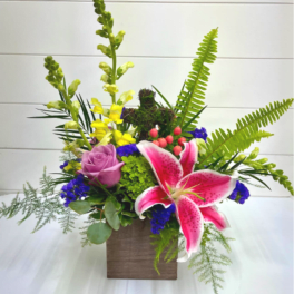 Mixed arrangement with pink lily, purple rose, yellow snapdragons, and ferns in a wooden cube.
