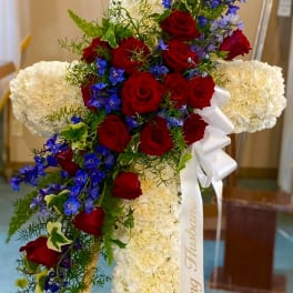 Cross-shaped floral tribute with red roses, white blooms, and blue flowers on an easel
