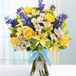 Bouquet of yellow roses, lilies, and blue flowers in a glass vase with a blue ribbon