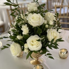White roses arranged in a gold vase on a table with small candles