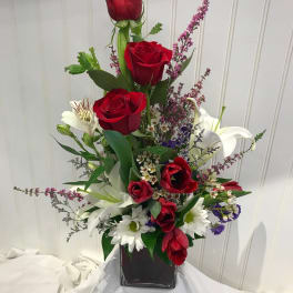 Tall arrangement of red roses, white lilies, and daisies in a square vase