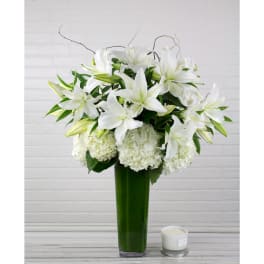 Tall arrangement of white lilies and hydrangeas in a green glass vase with a small white candle beside it.