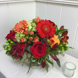Low round arrangement of red roses, gerbera daisies and mixed orange blooms in a clear glass bowl vase