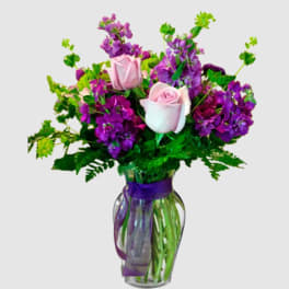 Pink roses and purple flowers arranged in a glass vase with a purple ribbon