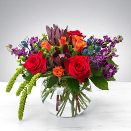 Mixed bouquet of red roses, orange blooms, and purple flowers in a clear glass vase