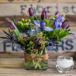 Purple tulips and blue irises in a square glass vase with a candle beside it