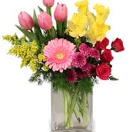 Mixed bouquet of pink tulips, yellow lilies, red roses, and pink gerbera daisies in a glass vase