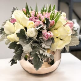 Low arrangement of ivory roses, pink tulips, and ranunculus in a round metallic bowl.