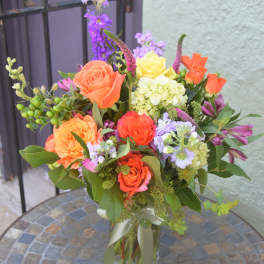 Mixed bouquet of orange roses, yellow blooms, and purple flowers in a clear glass vase