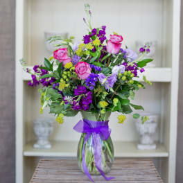 Mixed bouquet of pink roses and purple flowers with yellow accents in a clear vase tied with a purple bow