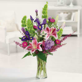 Tall mixed arrangement of pink lilies, purple blooms, and green accents in a clear glass vase