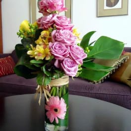 Tall glass vase with pink roses, yellow blooms, and a pink gerbera daisy framed by bold green leaves.