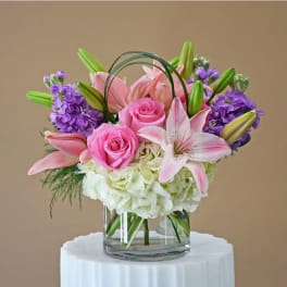 Round arrangement of pink roses and lilies with purple blooms in a clear glass vase