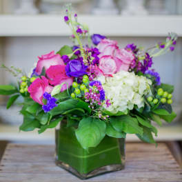 Low arrangement of pink roses, purple blooms, and white hydrangea in a square glass vase