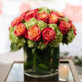 Low arrangement of orange and red roses with green hydrangeas in a clear glass vase beside a lit candle.