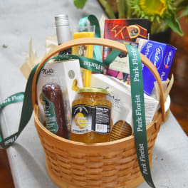 Gourmet food gift basket with marmalade, sausage, snacks, and a ribbon, with sunflowers in the background