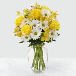 Yellow roses, white daisies, and yellow blooms arranged in a clear glass vase