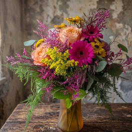 Bright mixed bouquet of peach roses, pink gerbera daisies, and yellow blooms in an amber glass vase