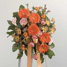 Tall standing spray of orange gerbera daisies and mixed flowers on an easel with peach ribbon.