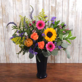 Tall mixed arrangement with sunflowers, pink gerbera daisies, and orange roses in a dark vase