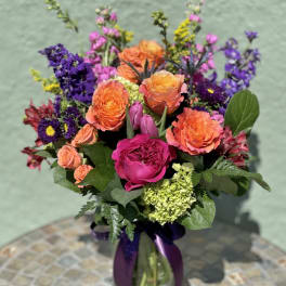 Bright mixed bouquet of orange roses, pink blooms, and purple flowers in a glass vase with a purple ribbon