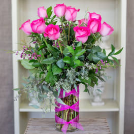 Tall arrangement of bright pink roses in a clear glass vase wrapped with pink ribbon