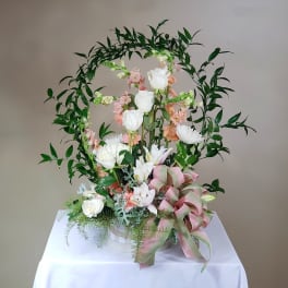 Floral arrangement with white roses and lilies in a basket, framed by greenery and a pink ribbon