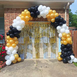 Black, white, and gold balloon arch framing a garage doorway with gold metallic fringe curtains.
