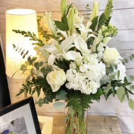Tall white lily, rose, and hydrangea arrangement with greenery in a clear glass vase on a wooden table