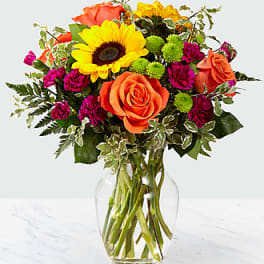 Mixed bouquet with sunflowers, orange roses, and magenta blooms in a glass vase