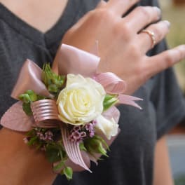 Wrist corsage with a white rose, pink ribbon, and small lavender flowers on a person’s arm