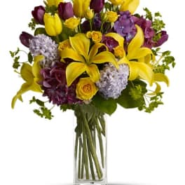 Yellow lilies and purple flowers arranged in a clear glass vase