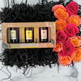 Boxed candle gift set on black shred with a row of pink and orange roses on the side
