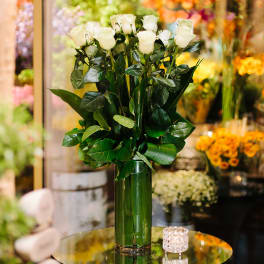 Tall arrangement of white roses with lush foliage in a green glass vase on a table