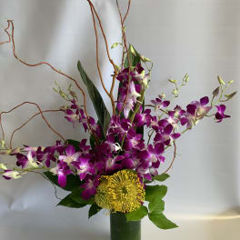 Purple orchids and a yellow pincushion bloom in a tall glass vase.