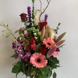 Tall mixed bouquet with pink gerberas, red roses, calla lilies, and orchids in a glass vase