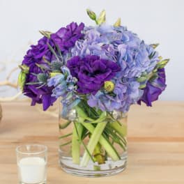 Purple and lavender flowers arranged in a clear glass vase beside a small candle